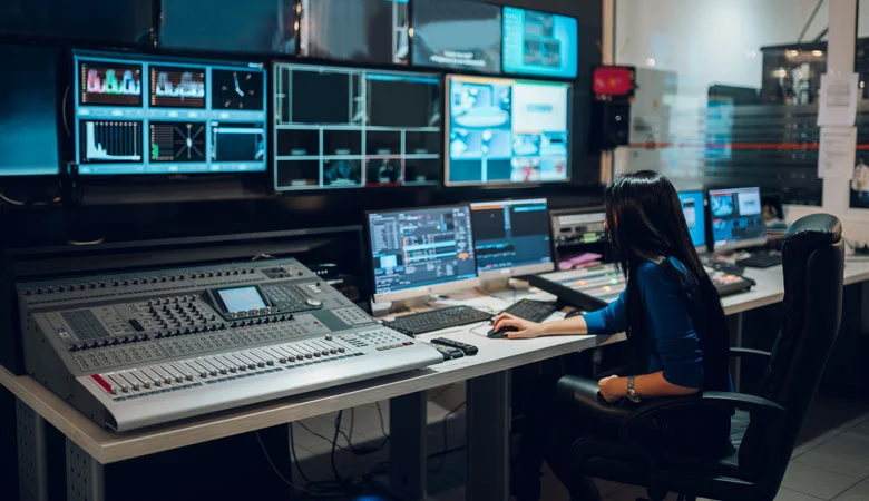 A broadcast engineer working inside a control room with audio mixer, multi view, automation system, graphics, video mixer, router control panel and intercom.