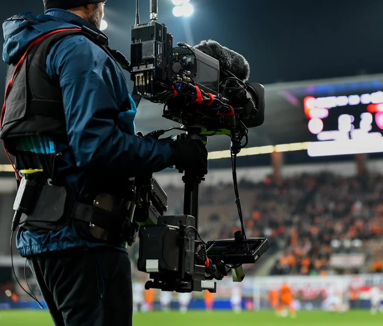 A broadcast cameraman covering a live sports event at a football stadium with a handheld Sony camera on a gimbal, part of a broadcast managed service.